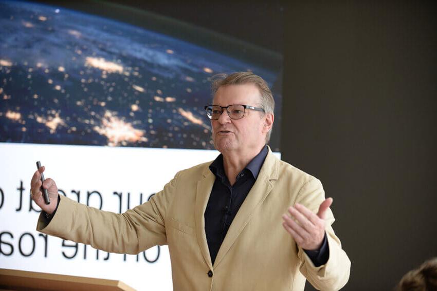 Antony Slumbers, in a tan suit and dark shirt, 在一场房地产优化活动中，他在幻灯片前做手势